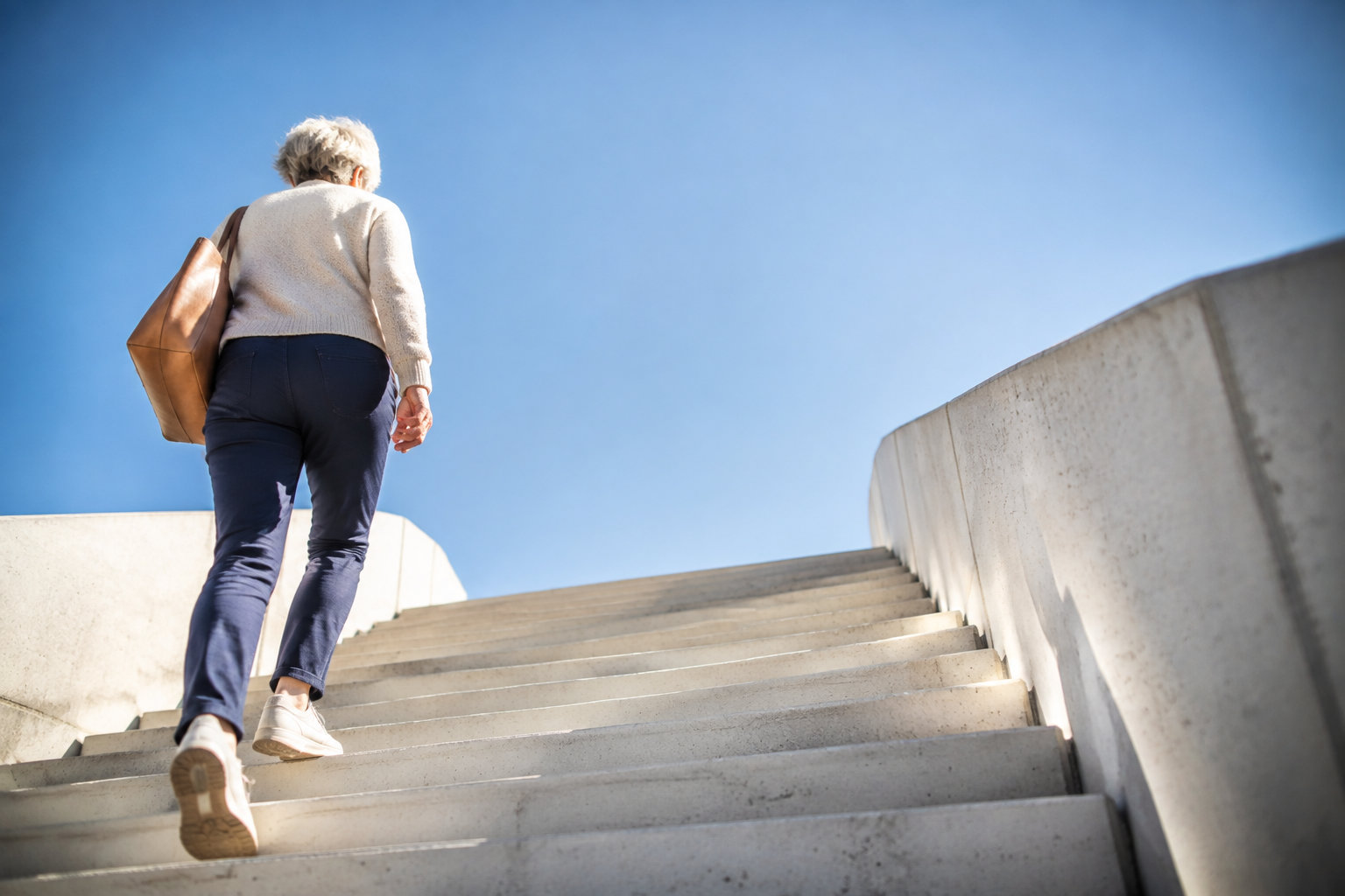 Une femme âgée monte des marches en béton vers un ciel bleu clair, symbolisant l'avenir et la transition.